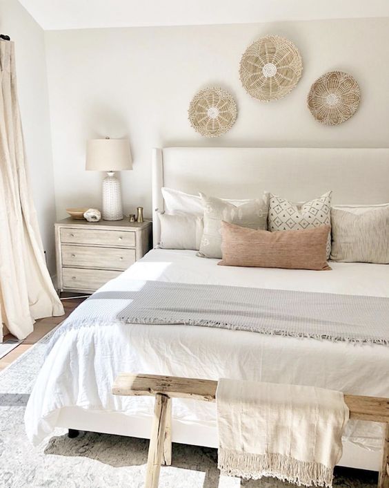 a neutral and warm bedroom with a creamy upholstered bed, a wooden bench, woven plates, printed pillows and neutral curtains