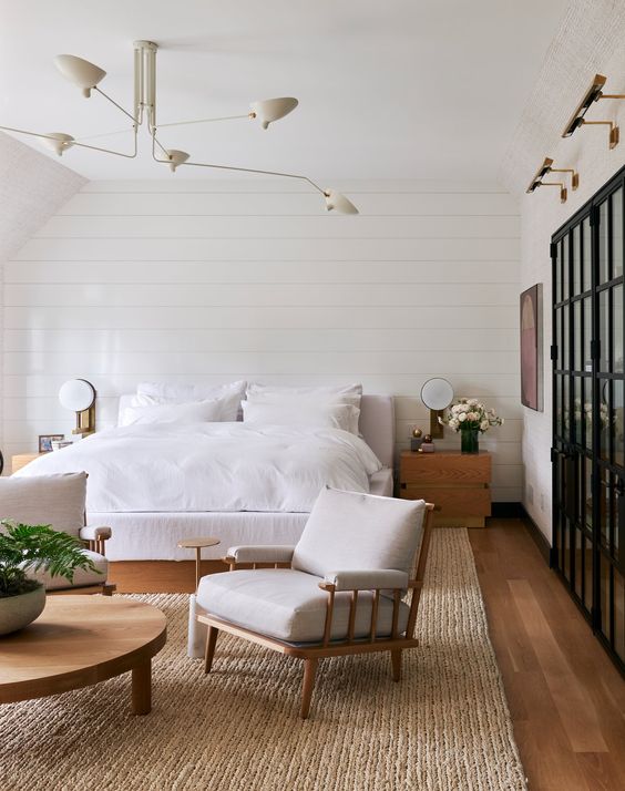 a neutral bedroom with white beadboard, a white bed and a chair, a wooden round table and some blooms and greenery