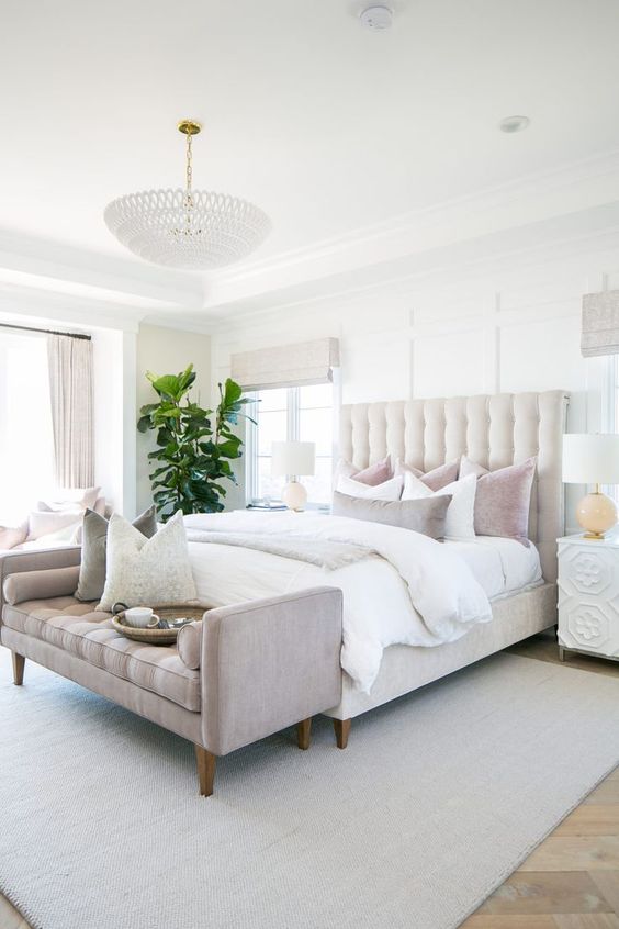 an elegant neutral bedroom with a creamy bed, neutral and pastel bedding, a pastel bench and a statement plant