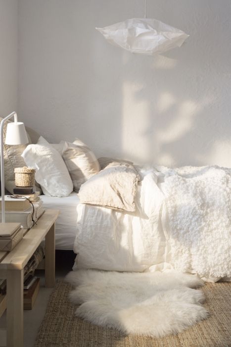 a neutral bedroom with white walls and layered bedding, faux fur, a wooden table with books and lamps instead of a nightstand