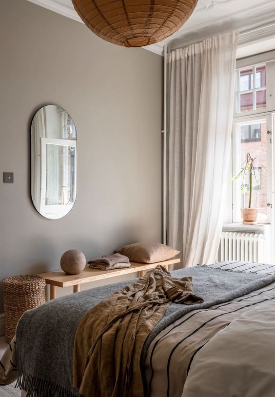 a beautiful greige bedroom with a bed with neutral and printed bedding, a wooden bench, warm-colored textiles, a paper pendant lamp and a basket for storage