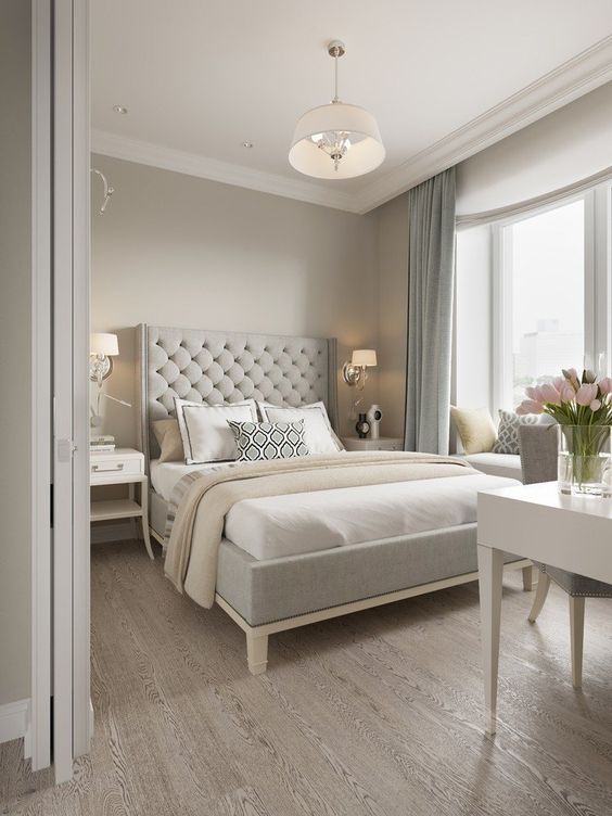 a chic greige bedroom with a grey upholstered bed, neutral bedding, a white vanity and a grey chair, blue curtains and some elegant lamps