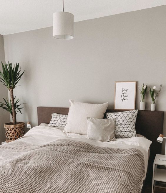 a cozy greige bedroom with a dark upholstered bed, potted plants and blooms, a white nightstand and printed bedding is inviting