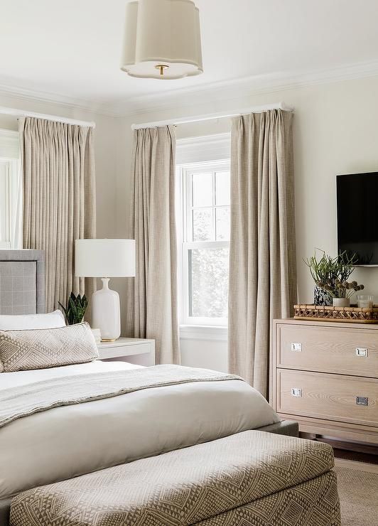 a greige bedroom with a grey upholstered bed, a stained dresser, beige curtains and an upholstered storage bench plus bedding