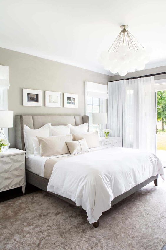 a greige farmhouse bedroom with a greige bed, neutral bedding, geometric nightstands, pendant and table lamps and a mini gallery wall