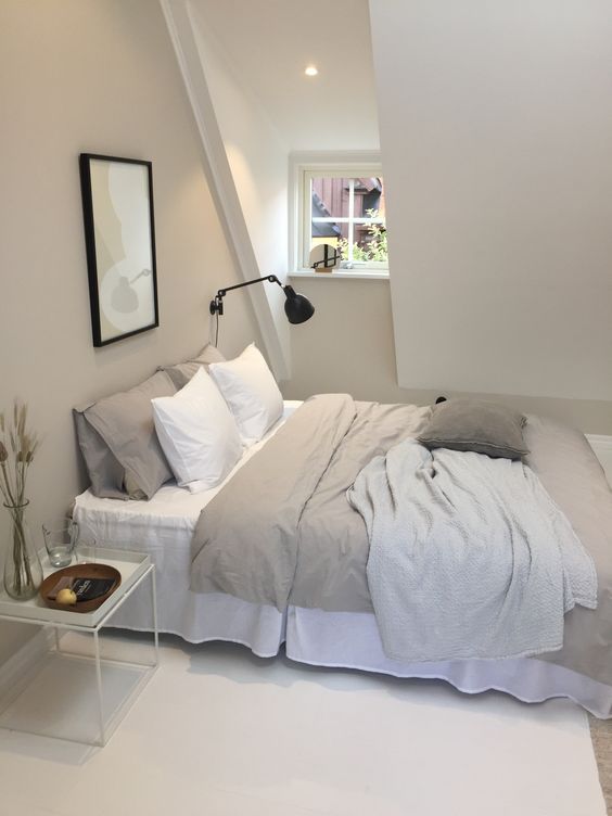 a modern greige attic bedroom with a bed, a nightstand and a black sconce, neutral and greige bedding, a small artwork is a cozy little sleeping space