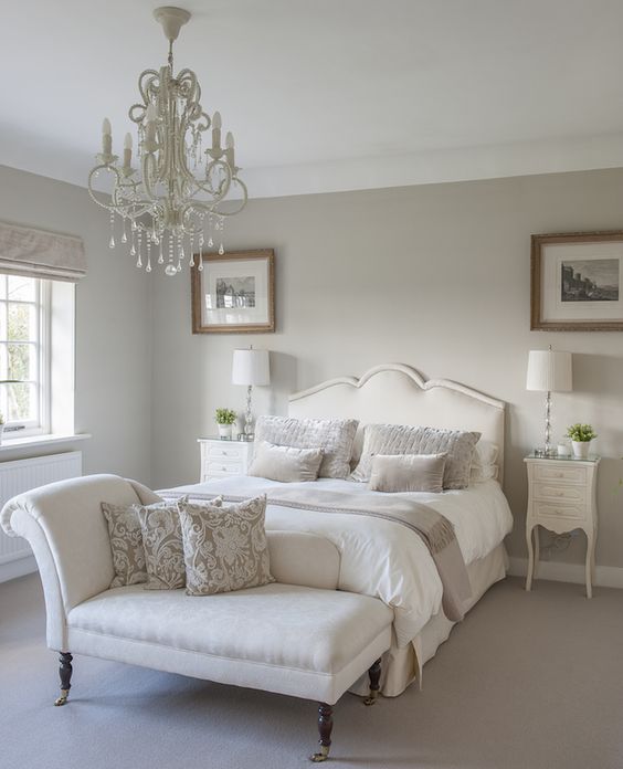 a sophisticated greige bedroom with creamy seating furniture and nightstands, an elegant white chandelier and small artworks