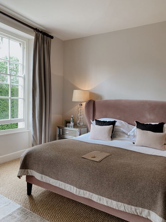 a stylish greige bedroom with a dusty pink velvet bed, neutral and blush bedding, mirrored nightstands, grey curtains and a jute rug