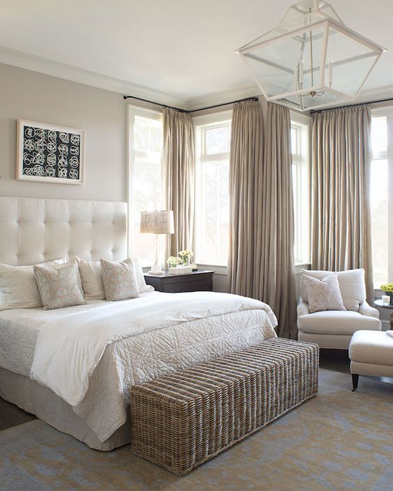 a welcoming greige bedroom with a creamy bed with a statement upholstered headboard, creamy seating furniture, a woven chest bench, greige curtains and a pendant lamp