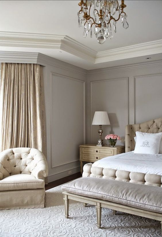 an exquisite greige bedroom with neutral upholstered furniture, a bench, neutral bedding, a crystal chandelier and tan printed curtains