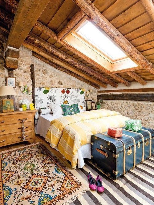 a catchy and colorful vintage attic bedroom with a floral bed, bold bedidng and rugs, a vintage blue chest and stone walls