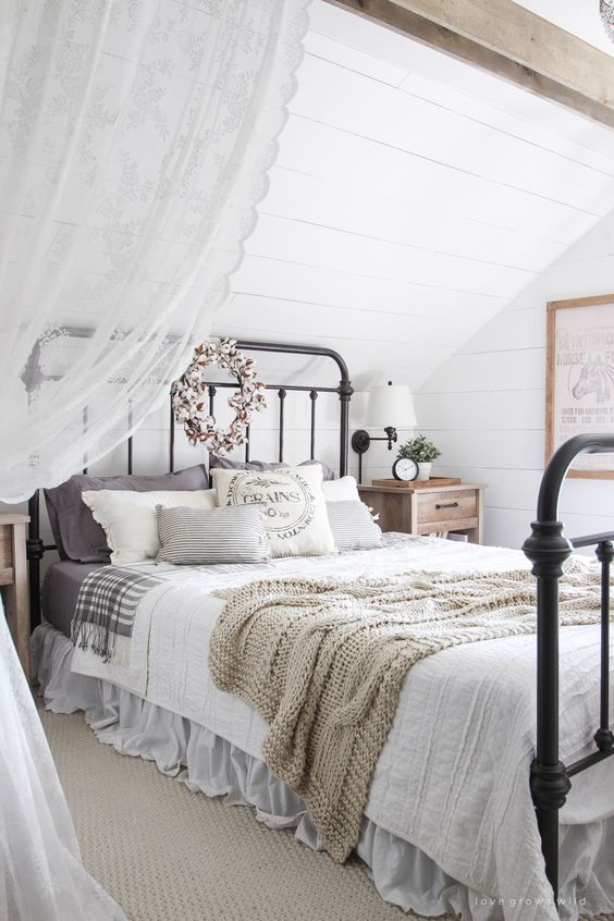 a neutral attic bedroom with a vintage and farmhouse feel, a metal bed, neutral and printed bedding, lace curtains and greenery