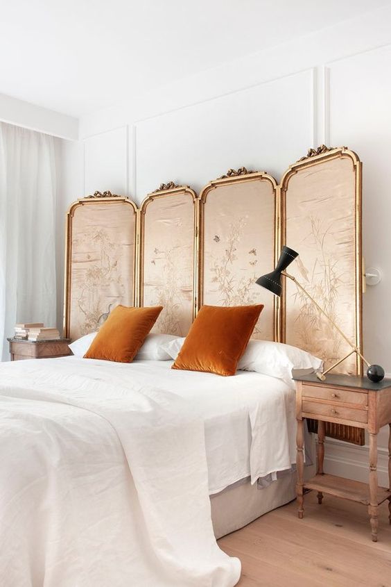 a refined vintage bedroom with a framed fabric screen, rust pillows, a statement lamp and wooden nightstands