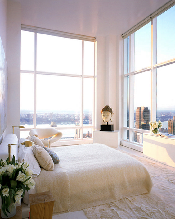 a stunning neutral bedroom with some cabinets, a bed, tree stumps, a bet chair and glass walls with the views of a big city