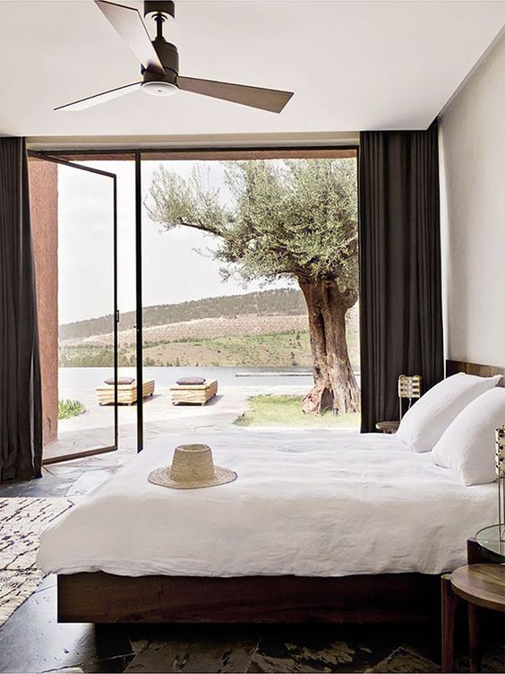 a relaxed modern bedroom with dark wooden furniture, neutral bedding and a glass wall with a door to the terrace plus black curtains