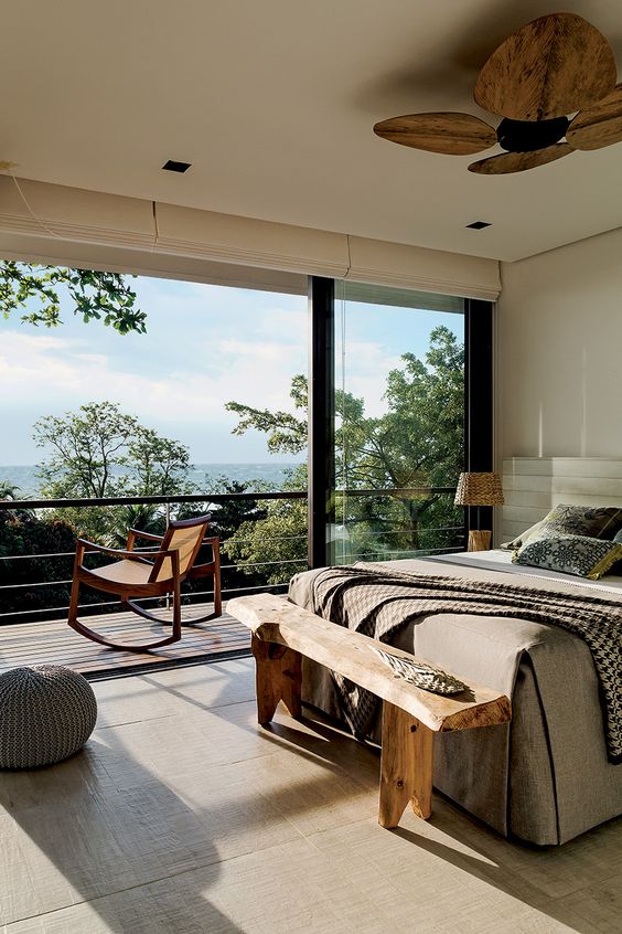 a cozy eclectic bedroom with an upholstered bed, a wooden bench and a fan, rattan lamps and a rocker chair on the balcony