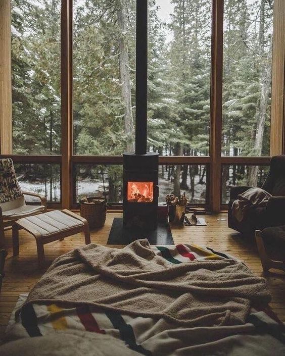 a cozy boho bedroom with a bed, comfy chairs and stools, a fireplace in the center and a glazed wall with a forest view