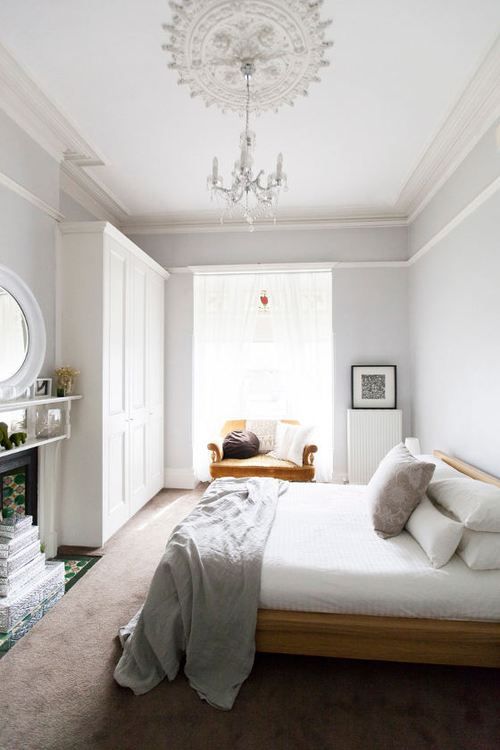 a neutral bedroom with white walls, molding on the ceiling, a wooden bed and a mustard chair, a fireplace and some books