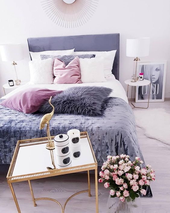 a cool feminine bedroom with a purple bed, a gold nightstand with candles, pink blooms, pink, white and purple pillows