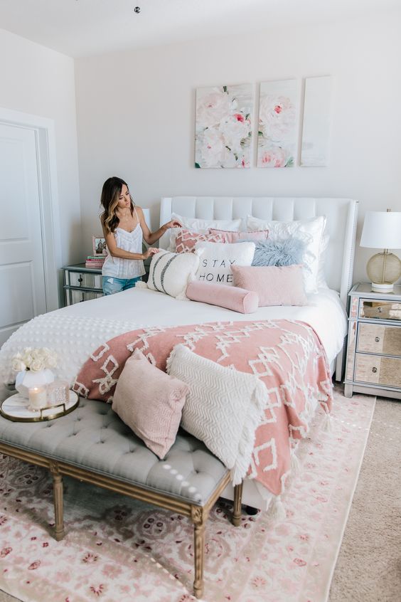 a modern feminine bedroom with a white upholstered bed, mirror nightstands, a grey bench, pink and white bedding and floral artworks