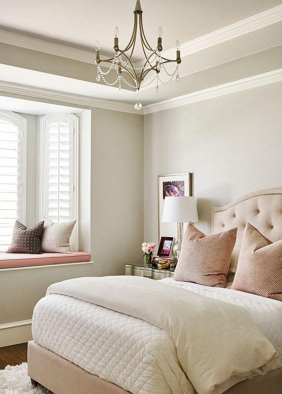 a neutral and pastel feminine bedroom with an upholstered bed, a crystal chandelier, blush and other pillows and a metallic nightstand