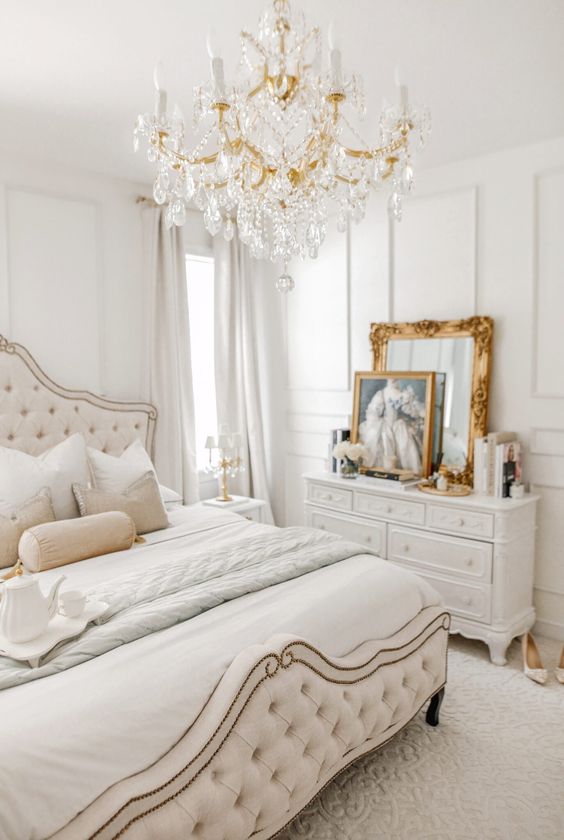 a neutral Versaille-inspired bedroom with refined furniture, a crystal chandelier, gold framed art and mirrors and layered bedding