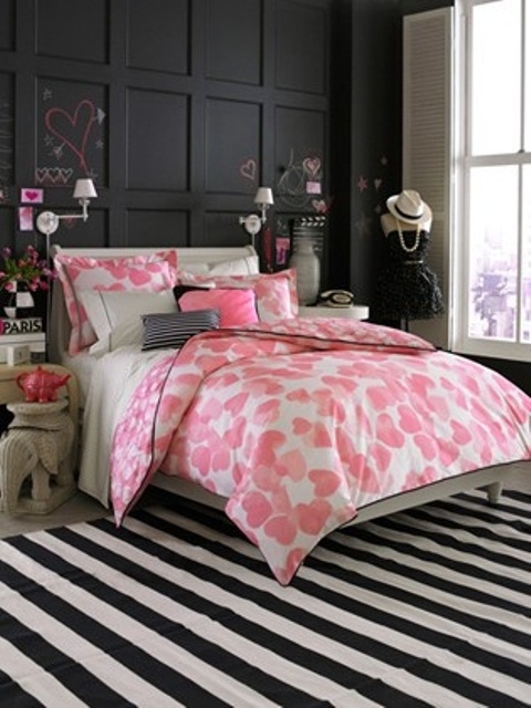 a bold feminine bedroom with a black paneled wall, neutral furniture, bright heart printed bedding, a striped rug and some more heart decor