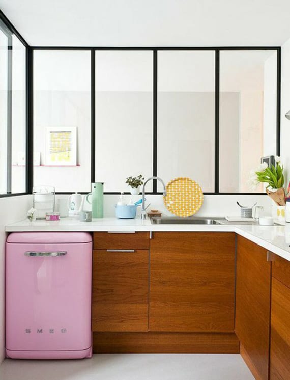 small corner kitchen in the living room with glazing, white corner worktop over wooden kitchen cabinets and a small retro refrigerator in pink